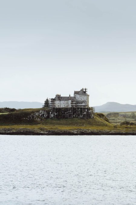 Duart Castle on its rocky headland across calm water, wrapped in scaffolding for repairs in 2021, hills behind.