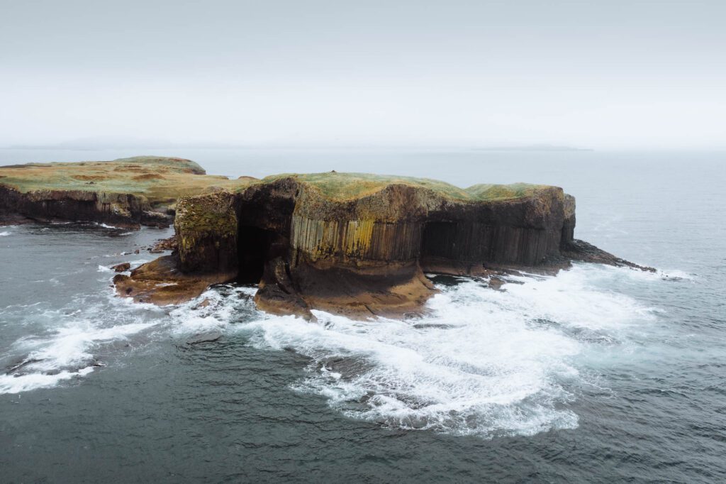 Basalt columns and sea caves on a grassy headland surrounded by breaking waves