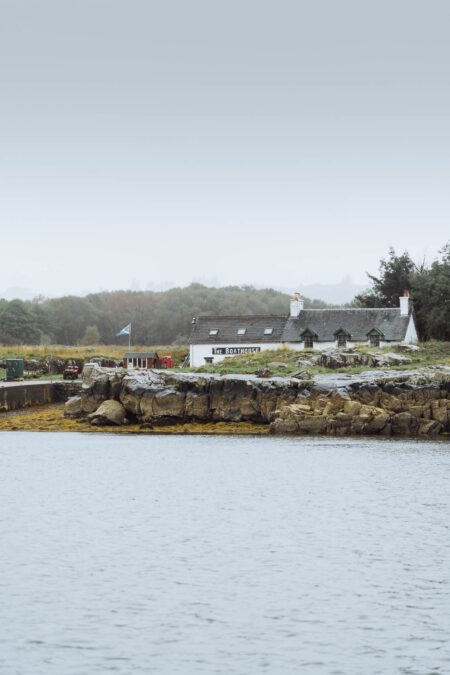 The Boathouse on Ulva across the sound, white on the rocky shore with a Saltire flying.