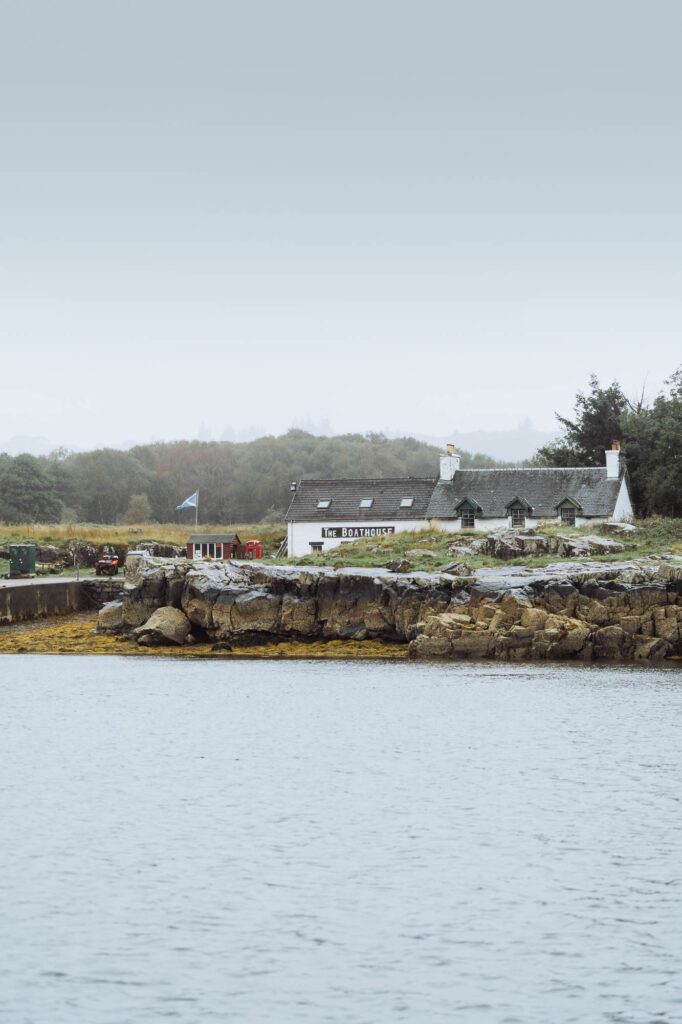 The Boathouse on Ulva across the sound, white on the rocky shore with a Saltire flying.