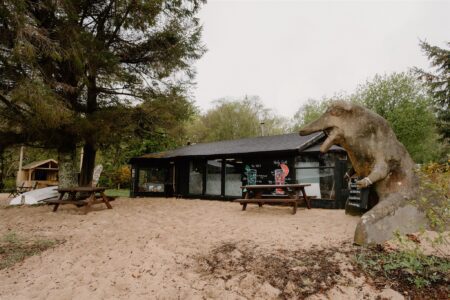 Wooden dinosaur sculpture beside a black timber café with picnic tables on sand