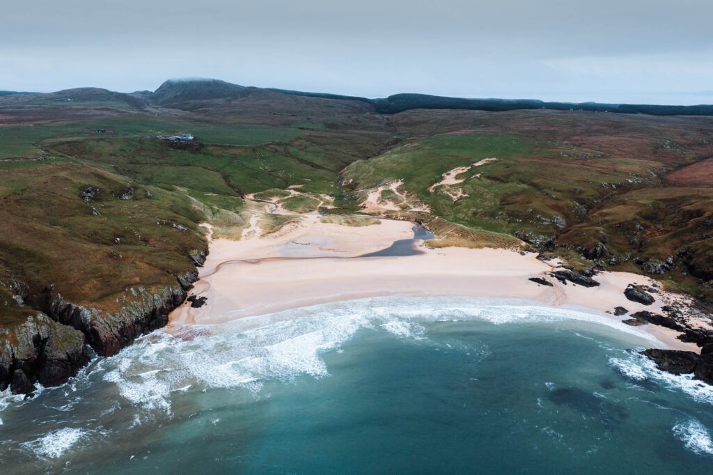 Lossit Bay from above, a crescent of pale sand between rocky headlands, surf rolling in.