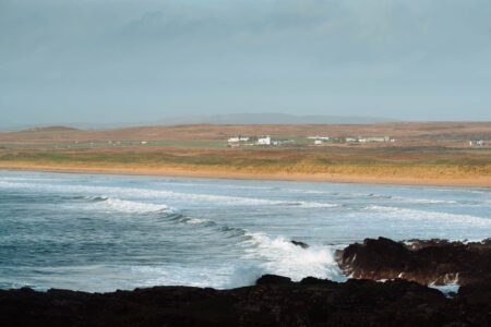 Surf breaking on dark rocks at Laggan Bay, the dunes and white farmhouses beyond.