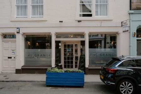 White-fronted shopfront reading "The Grange", with a blue planter and parked car outside