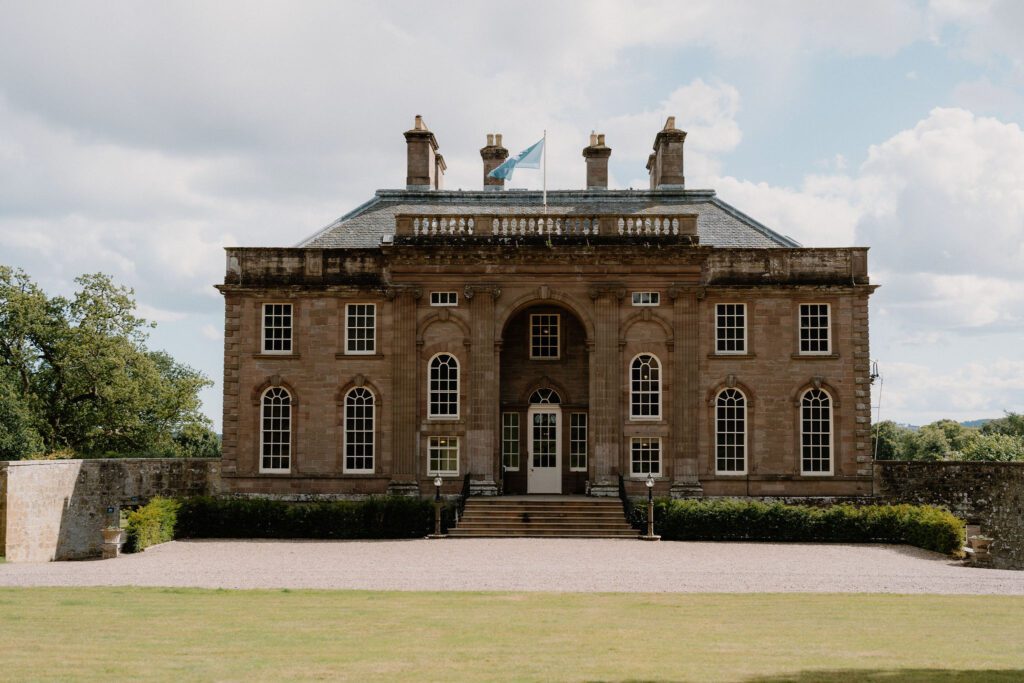 Sandstone mansion with columned entrance, chimneys and flag against a cloudy sky