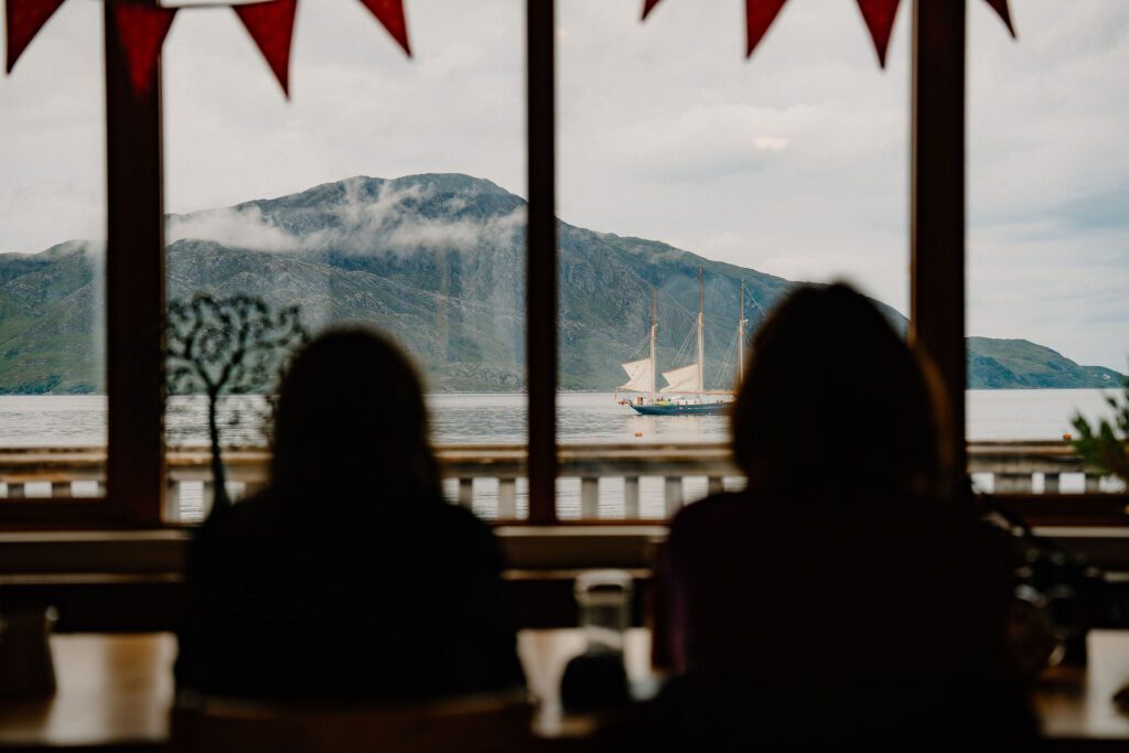 Two silhouetted diners watch a tall ship sail past mountains beyond the window
