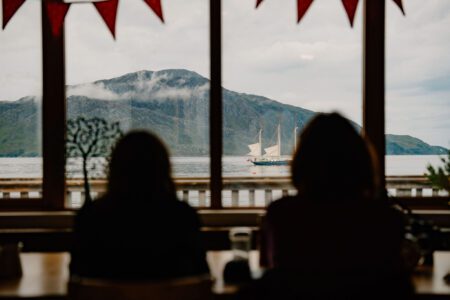 Two silhouetted diners watch a tall ship sail past mountains beyond the window