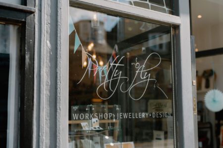 Shop window lettered "Pretty Fly, Workshop, Jewellery, Design" with bunting reflected inside