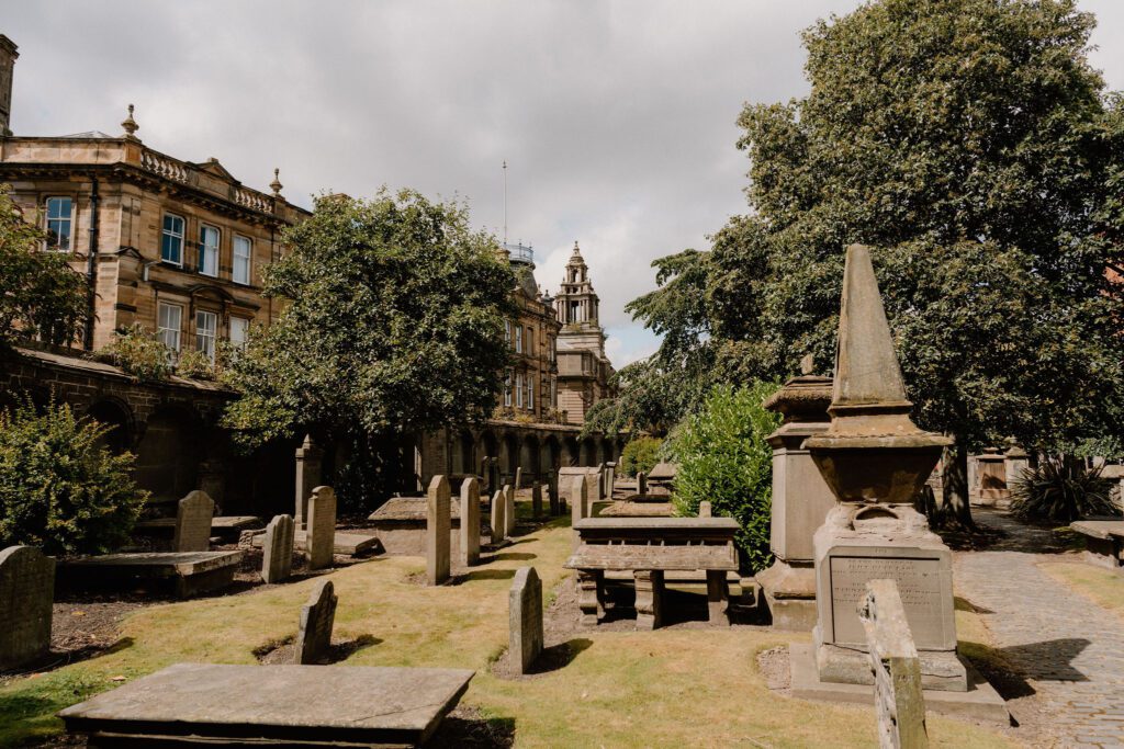Gravestones and an obelisk monument among trees beside sandstone buildings