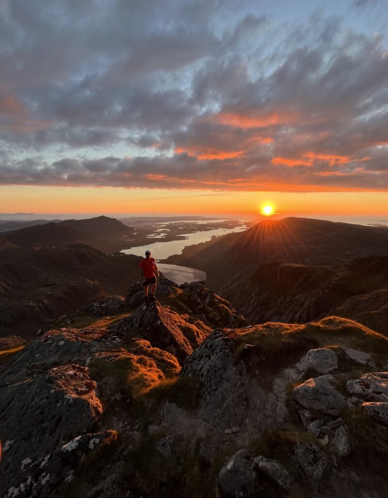 Hiker in red watches the sun set over a mountain ridge