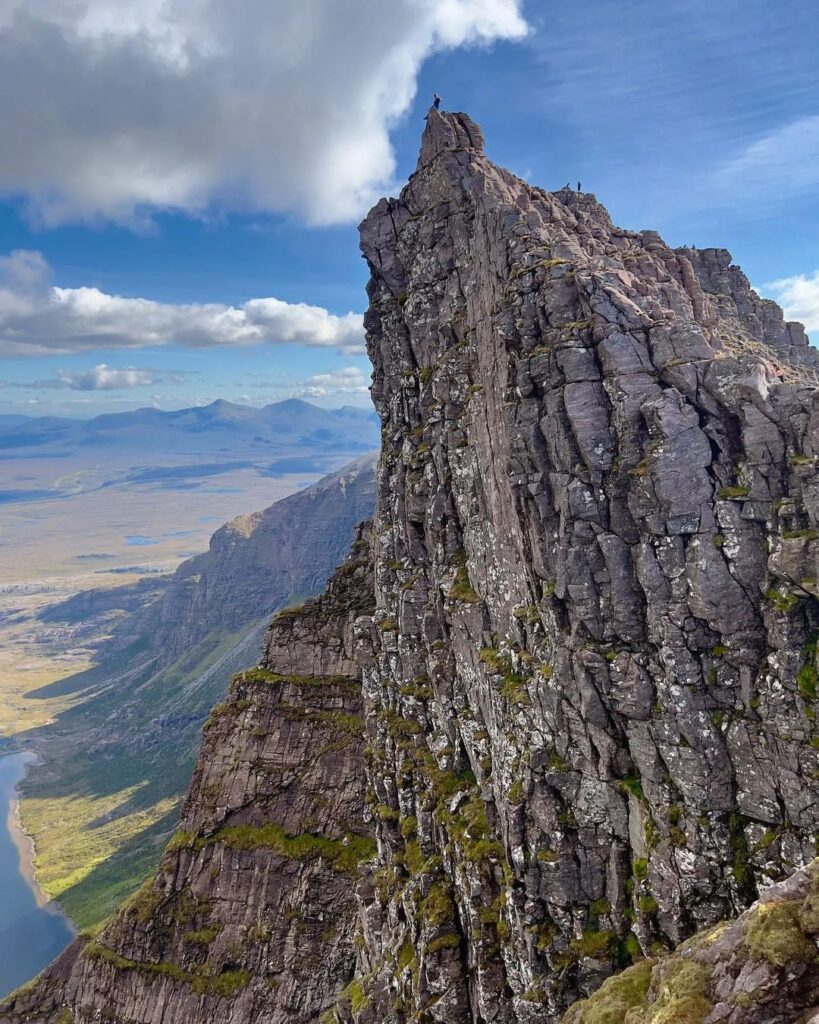 A climber stands atop a jagged rock pinnacle above a glen and loch