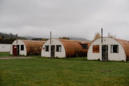 Rusted Nissen huts with white gables stand in a grass field under grey sky