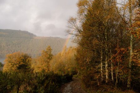 Faint rainbow over a forested loch, path lined with autumn trees