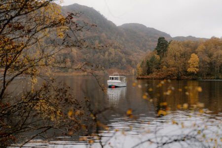 Small boat crossing a loch, framed by autumn branches and hillside trees
