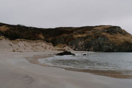 Sandy beach curves beneath grassy cliffs beside a rocky outcrop in the sea