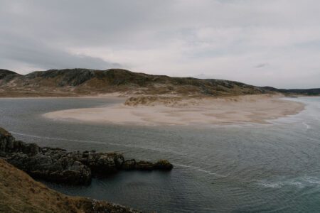 Sandy beach curves between hills and rocky shoreline under grey skies