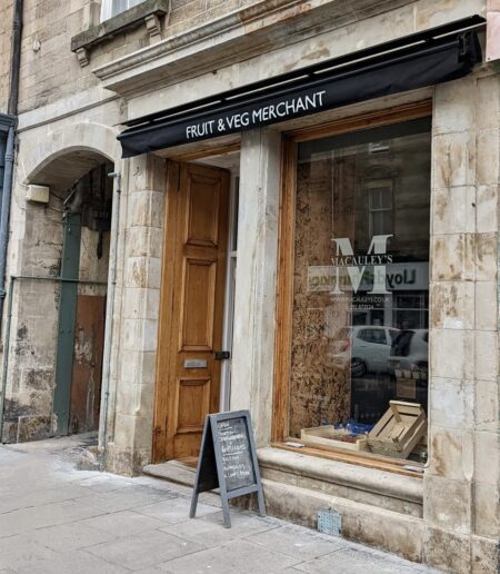 Wooden shopfront with black awning reading "Fruit & Veg Merchant" and Macauley's signage in the window
