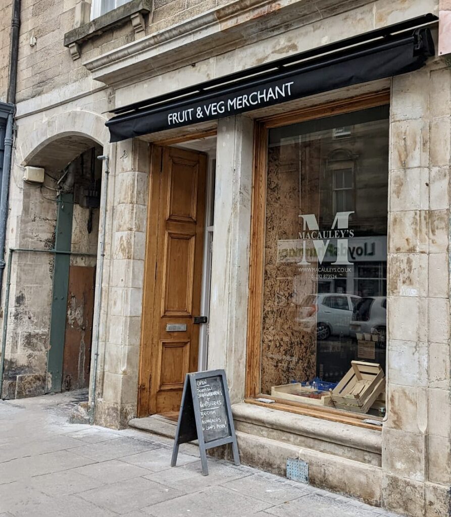 Wooden shopfront with black awning reading "Fruit & Veg Merchant" and Macauley's signage in the window