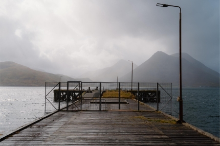 Wooden pier with metal railings leads towards mountains under cloudy skies