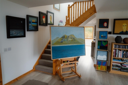 The stair hall of Raasay Gallery hung with paintings to the ceiling, a large canvas of a green island standing on an easel.