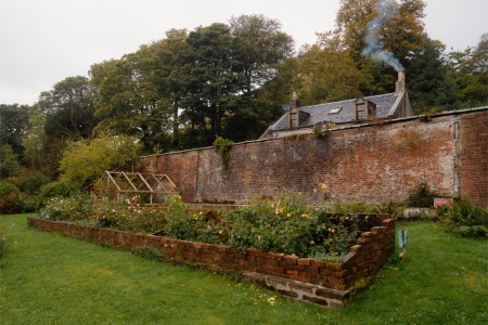 Brick-walled garden with raised beds and a cold frame, smoking chimney beyond