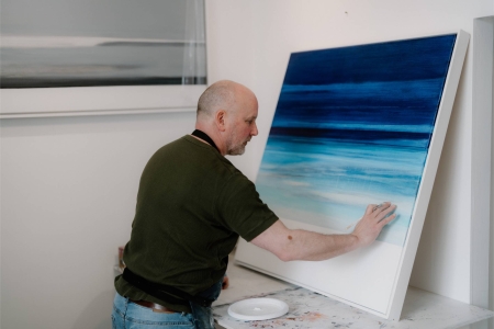 Artist touching a large blue seascape painting propped against a studio wall