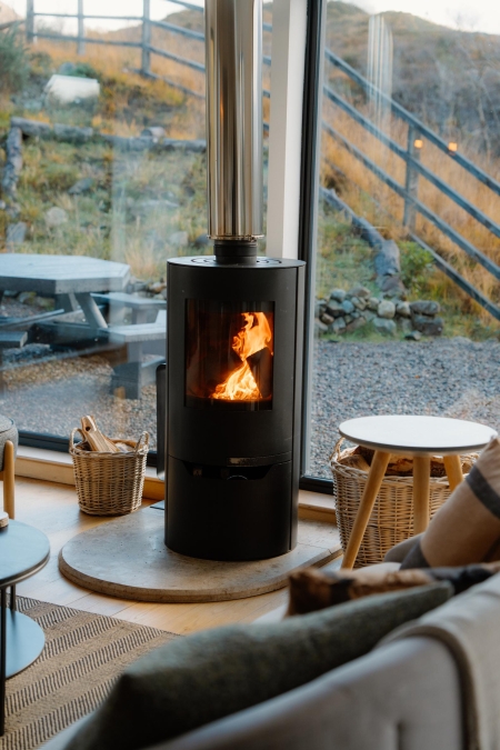 Wood-burning stove alight beside floor-to-ceiling windows overlooking a hillside path