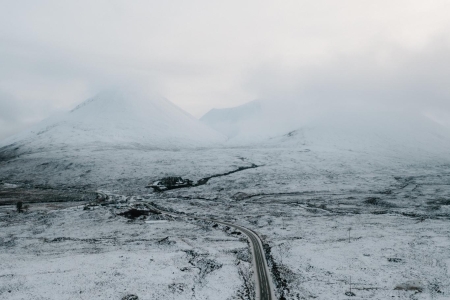 Snow-covered road winding towards mist-shrouded mountains under a grey sky