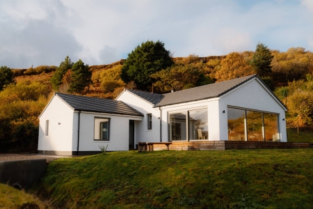 A white bungalow with a glazed gable end sits before an autumn hillside, a wooden bench on the lawn in front.