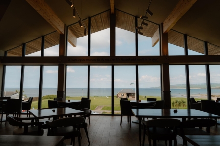 Timber-framed dining room with tall windows overlooking a grassy shoreline and sea