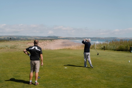 Two golfers on a coastal tee, one mid-swing overlooking a sandy bay