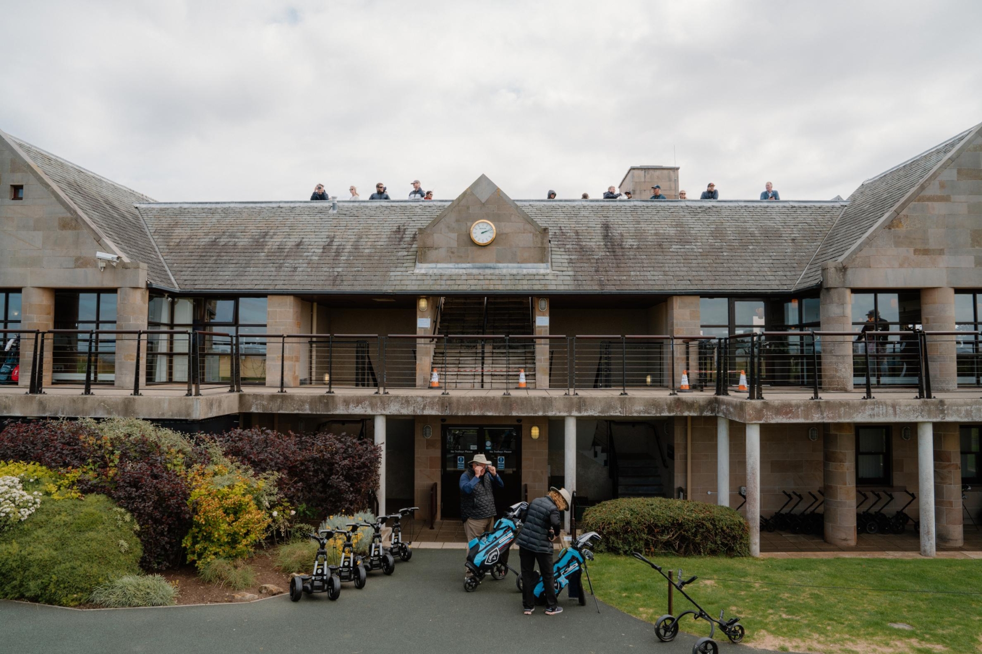 Golfers with trolleys outside a clubhouse, spectators lining the roof terrace above