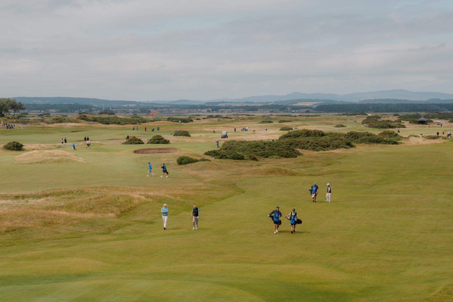Golfers and caddies walk the fairway on a links course under grey skies