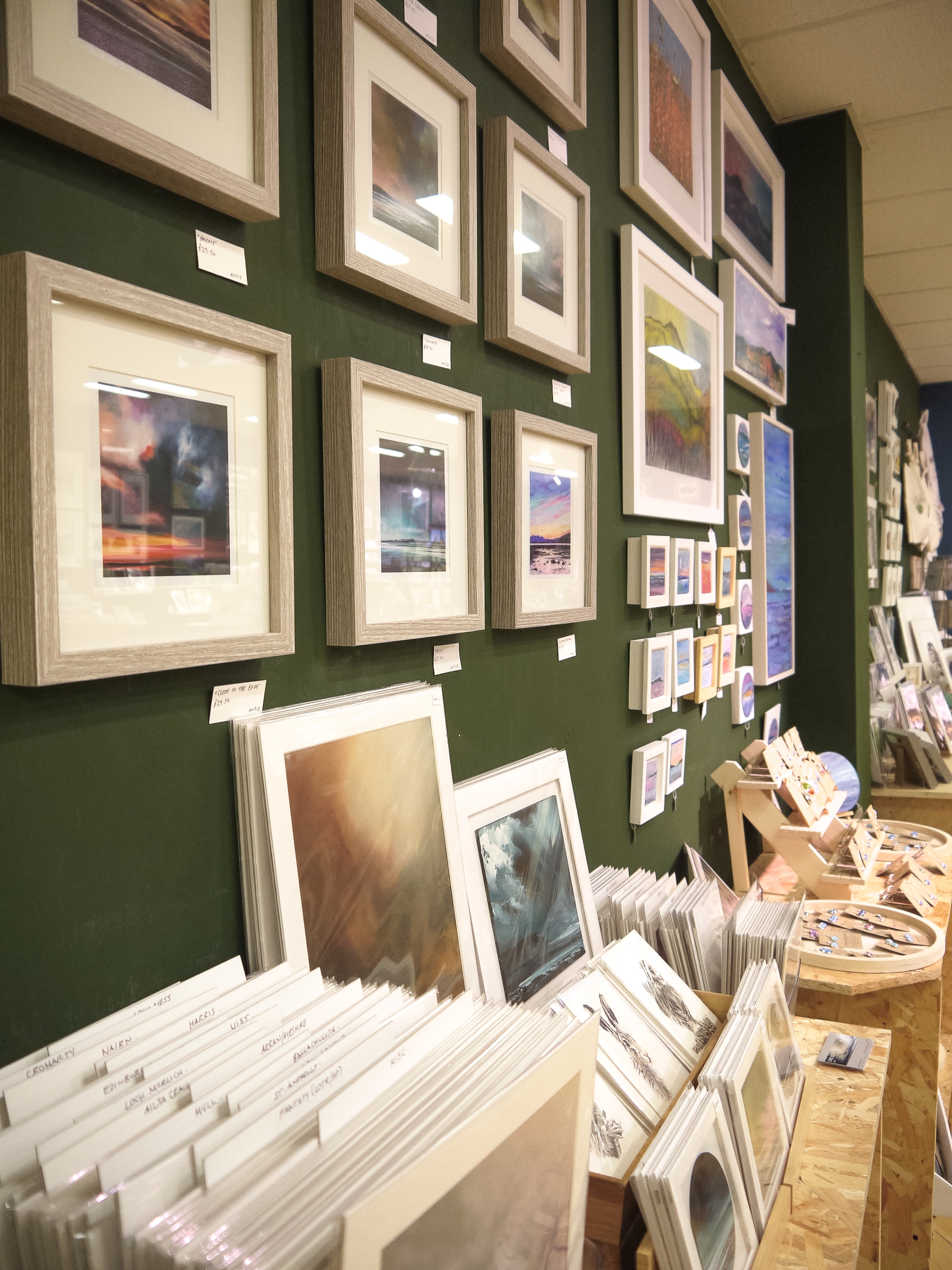 Framed prints on a dark green wall above racks of cards at Made in Stirling.