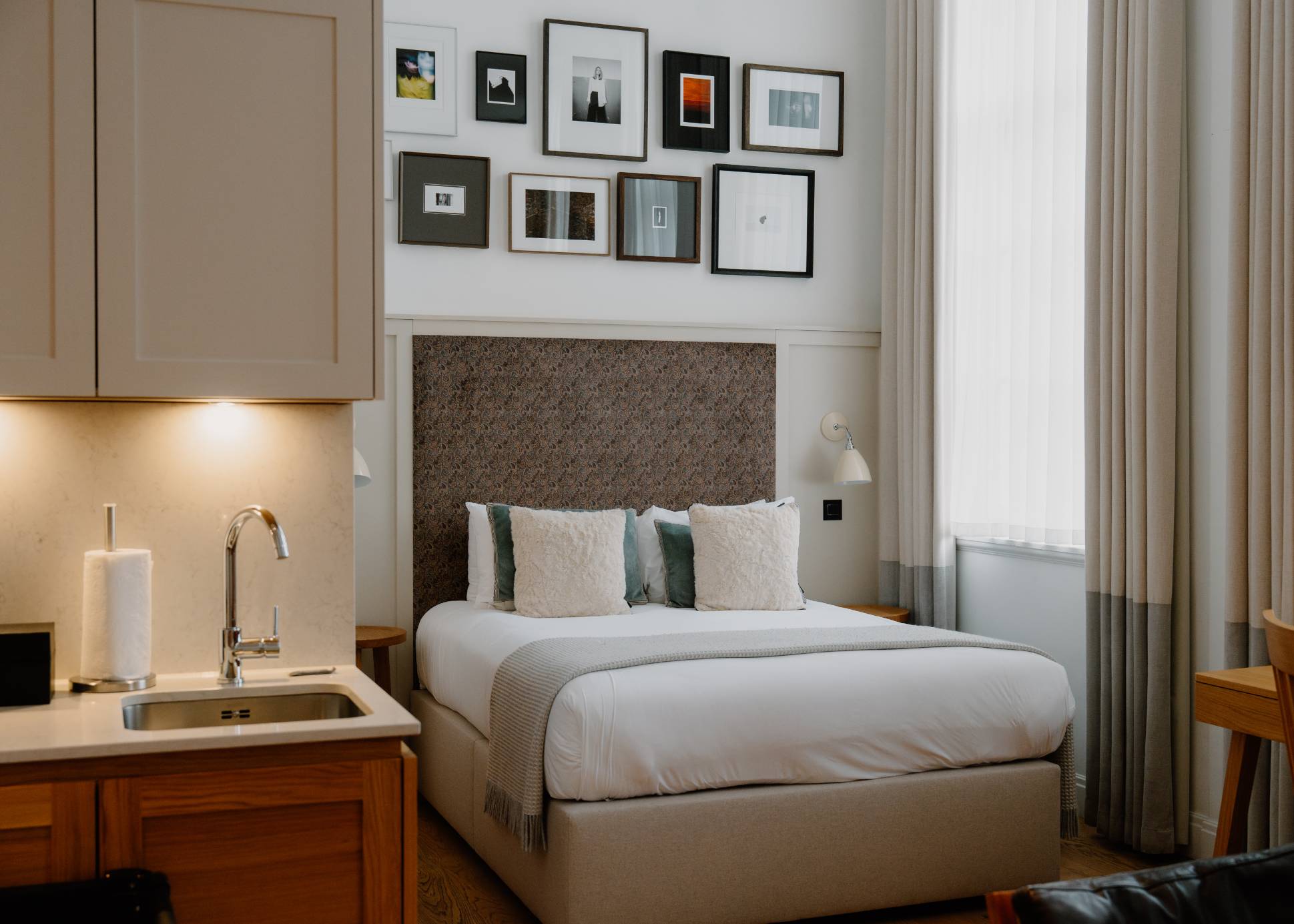 A studio bedroom with a kitchenette sink and cream cabinets beside a bed under a wall of framed photographs, tall curtains at the window.