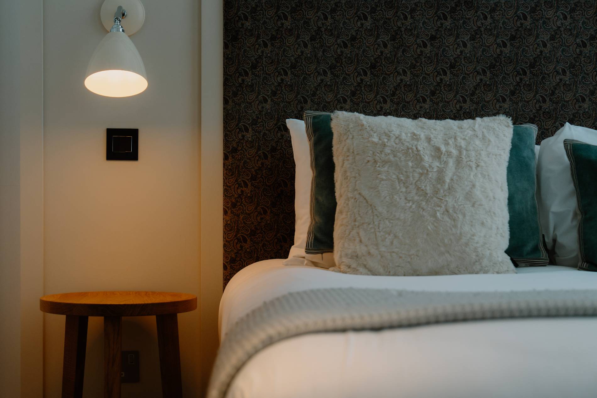 A dome wall lamp lights a round wooden stool beside a bed with a dark patterned headboard, a cream textured cushion and teal-edged pillows.