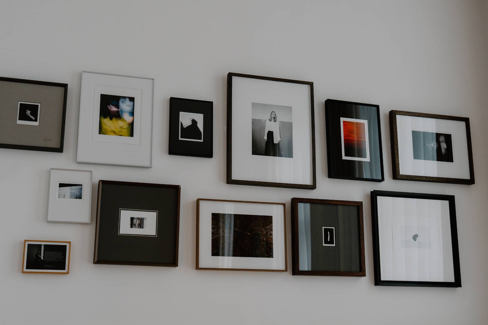 A grid of small framed photographs and prints in black, white and wooden frames hangs across a plain wall.