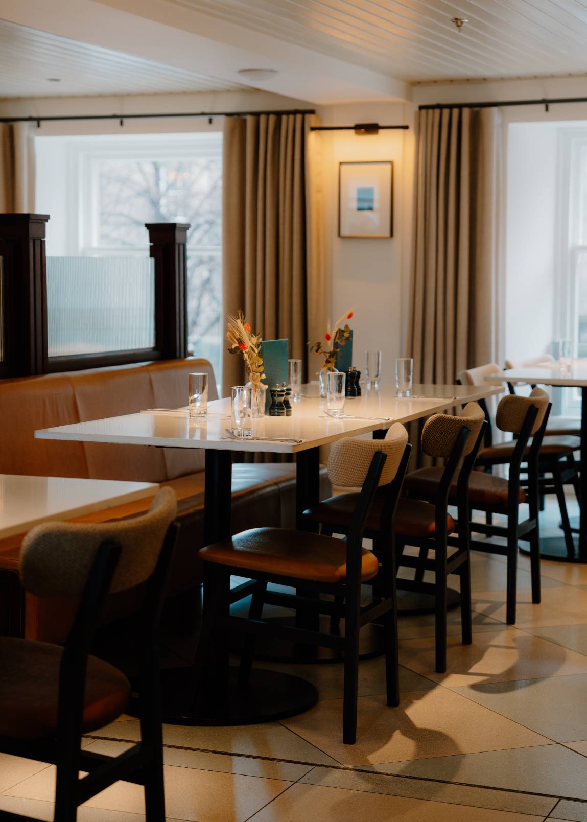 A long dining table set with candles and flowers stands before curtained windows, dark chairs arranged along both sides on a tiled floor.