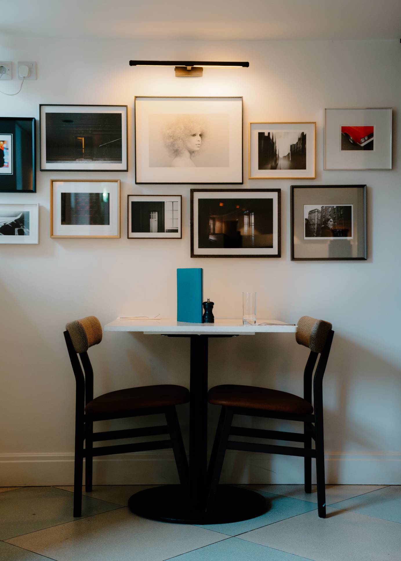 A small dining table set with a blue menu and glasses stands below a gallery of framed photographs, two chairs with woven backs on either side.