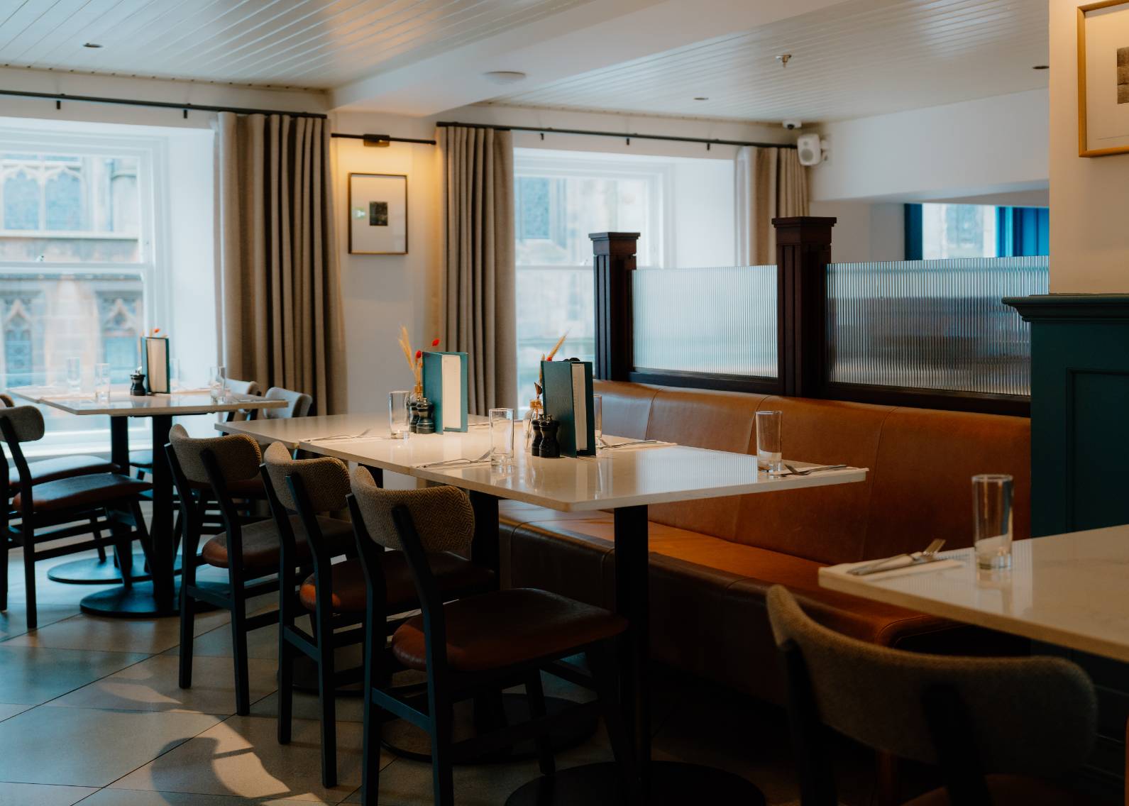 A dining area with dark leather banquette seating and chairs stands before curtained windows, framed pictures on the wall beside frosted glass panels.