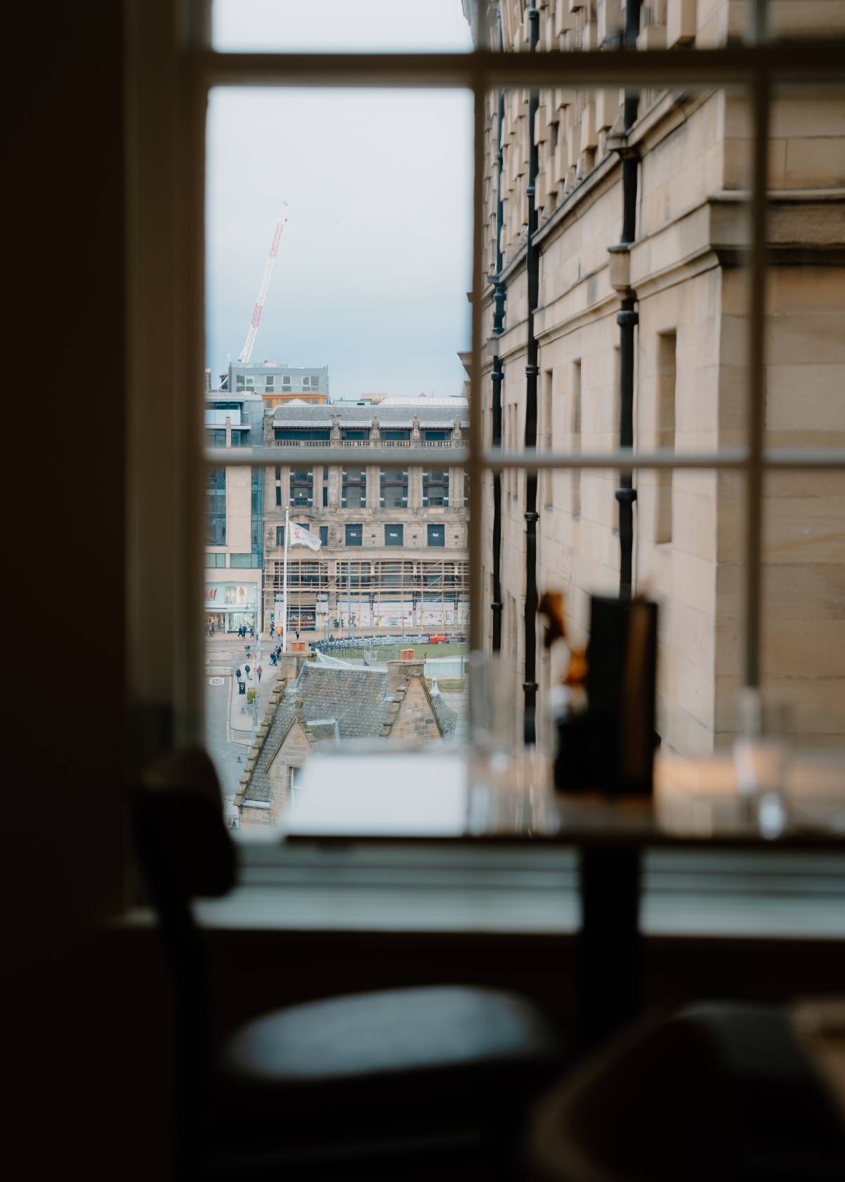 A crane and rooftops are seen through a paned window, a table with glasses in the foreground of a dimly lit dining room.