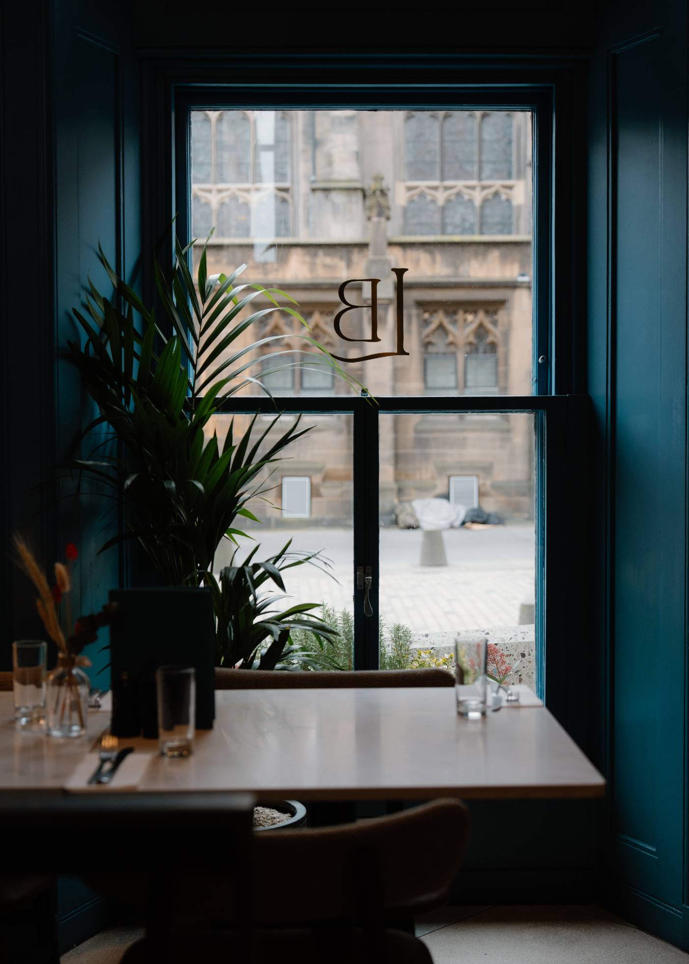 A monogrammed window looks onto a church facade, potted plants and a dining table set with glasses in the foreground of a teal-panelled room.