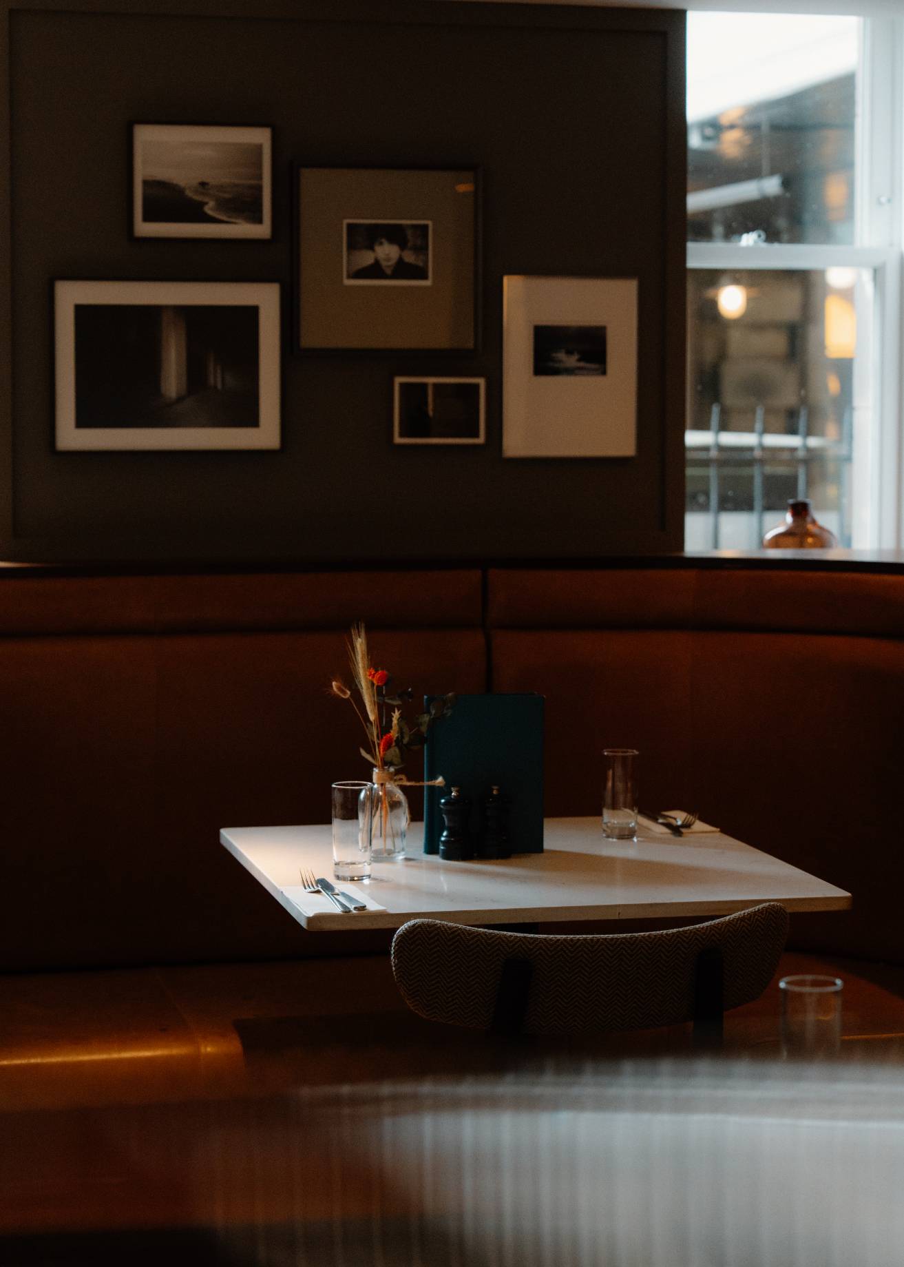 A gallery of black and white photographs hangs above a leather banquette, a vase of dried flowers and a menu set on the white table in front.