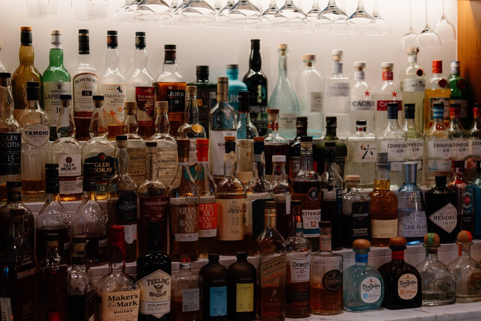Rows of labelled spirit bottles line a bar shelf, wine glasses hanging above.