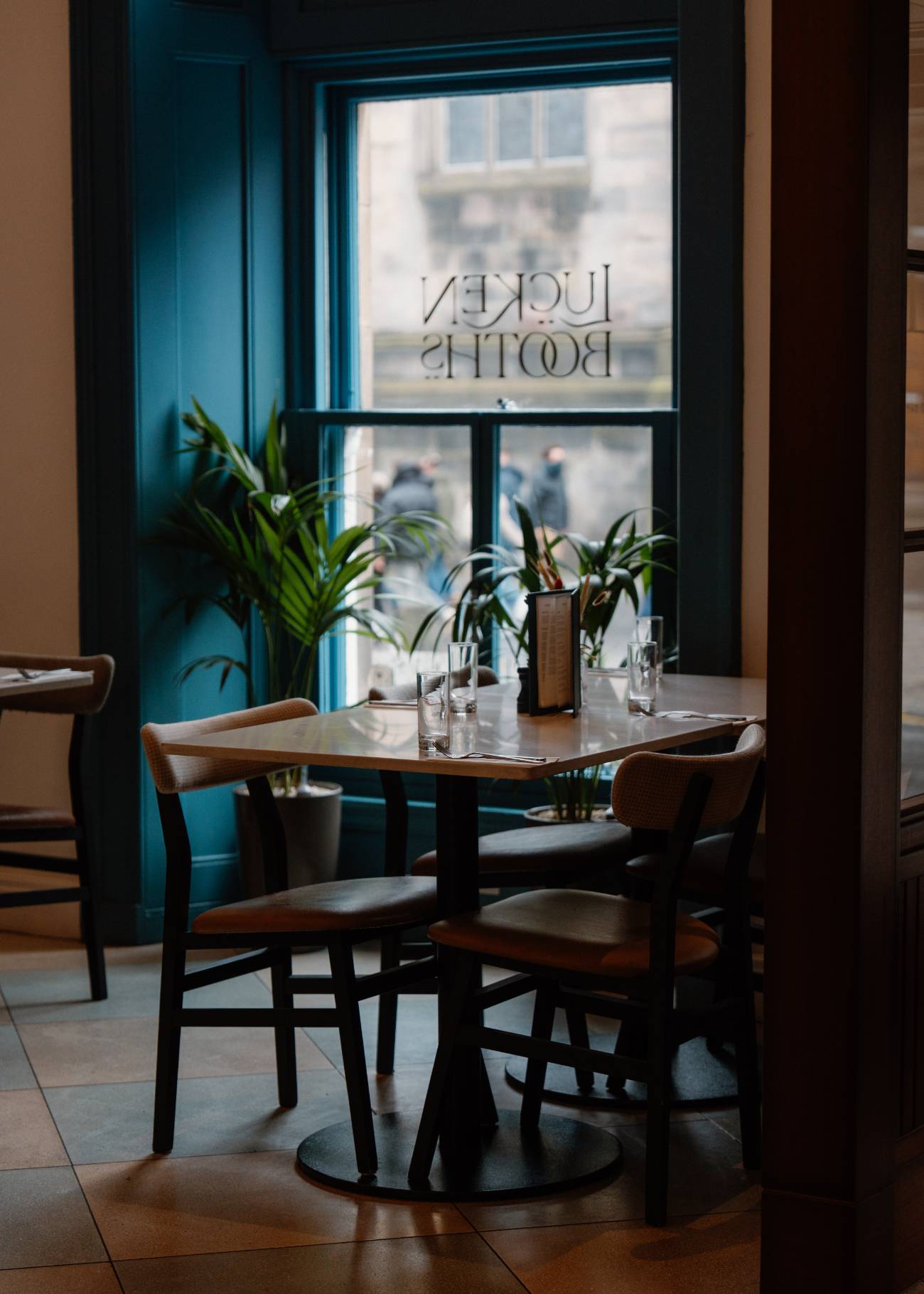 A dining table set with glasses stands before a window etched with the restaurant's name, potted plants and dark wooden chairs around it.