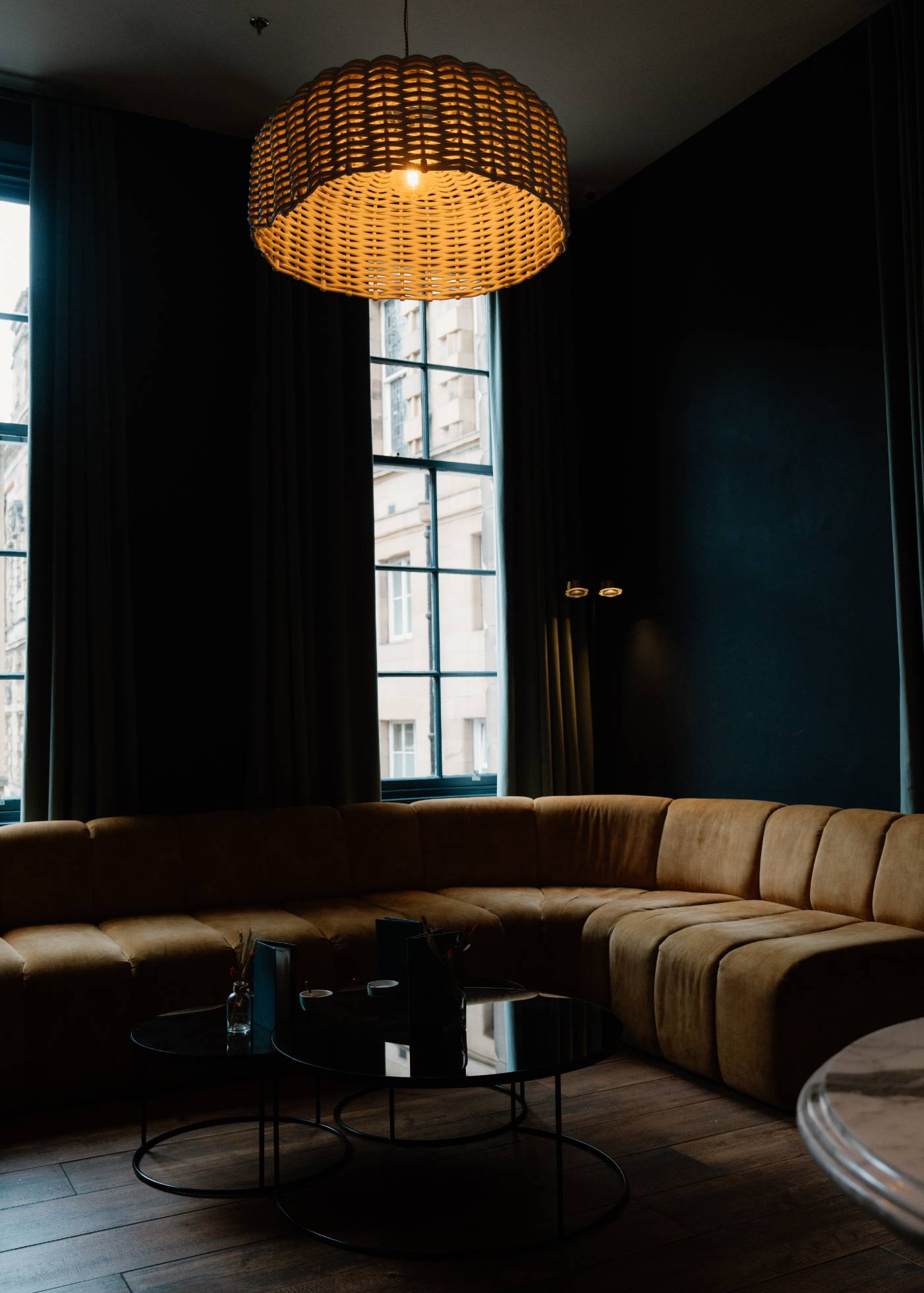 A wicker pendant light hangs above a curved mustard velvet sofa, round black coffee tables in front and tall windows behind.