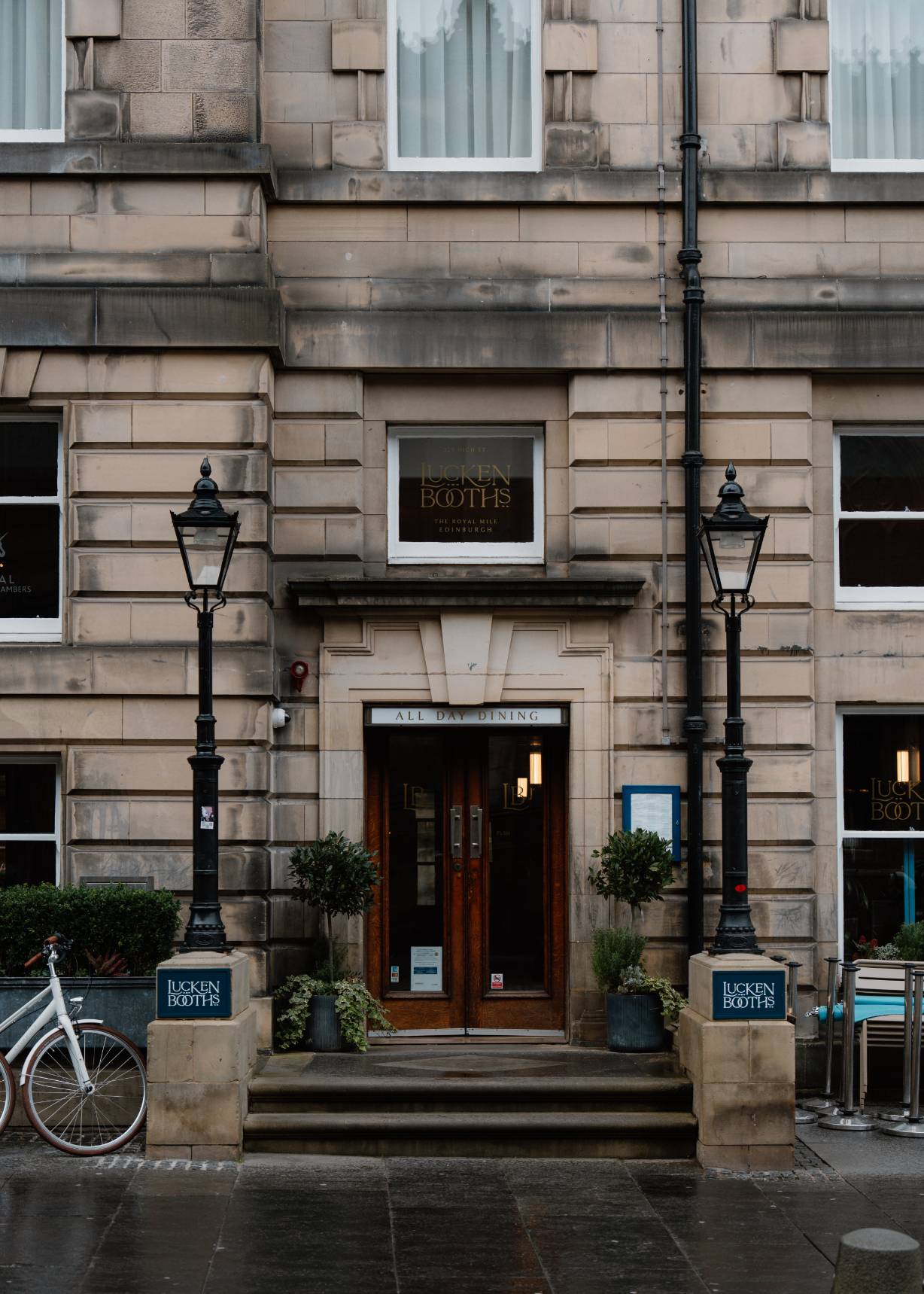 A restaurant entrance reads "Lucken Booths, All Day Dining" beneath an arched stone facade, lanterns and topiary either side of the door.