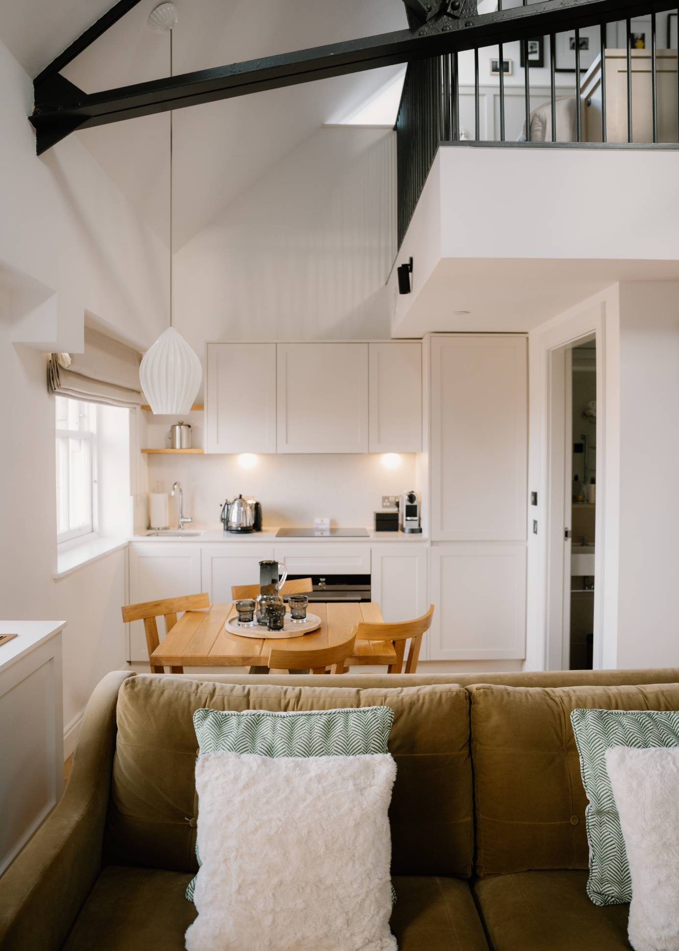 A galley kitchen with white cabinetry leads to a dining table and a mustard sofa with cushions, a mezzanine landing visible above.