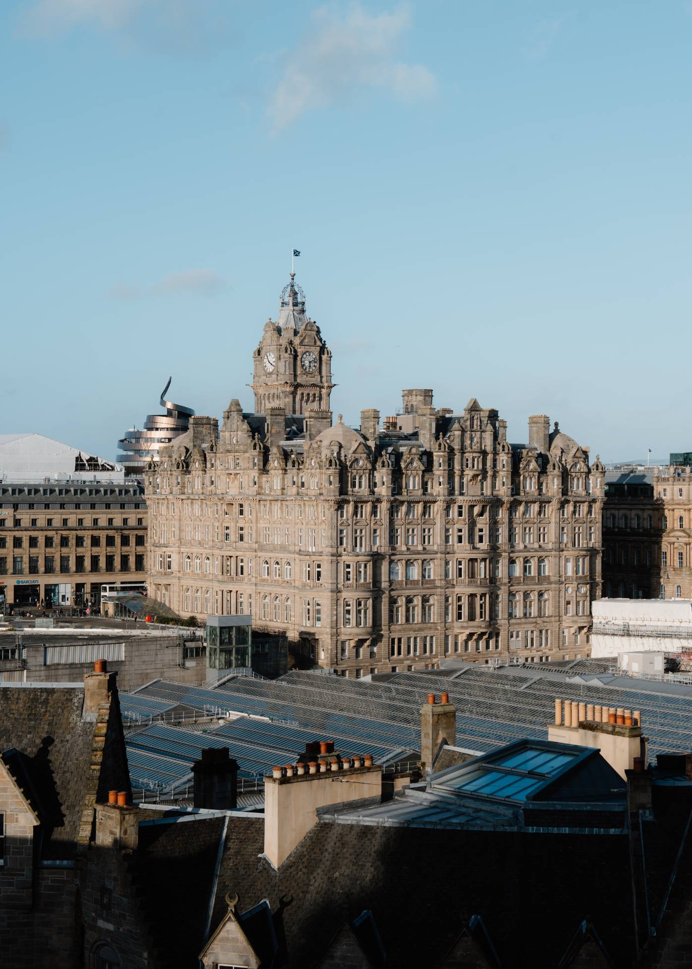 An aerial view of Edinburgh shows the Balmoral Hotel clock tower among rooftops and chimneys under a clear sky.