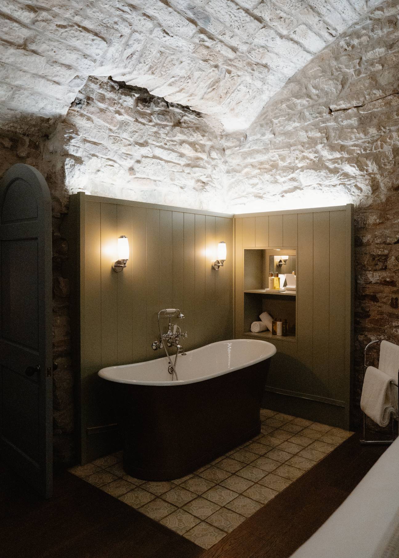 A roll-top bath with a dark exterior sits under a vaulted stone ceiling, wall lights either side and a shelf of toiletries in an alcove.